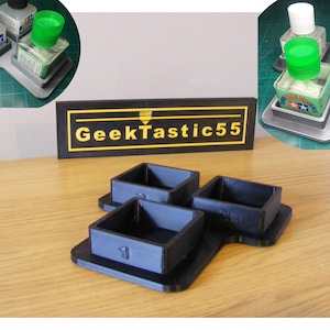 May include: A 3D printed organizer for paint bottles with three square compartments. The organizer is black and has a GeekTastic55 logo on the front.