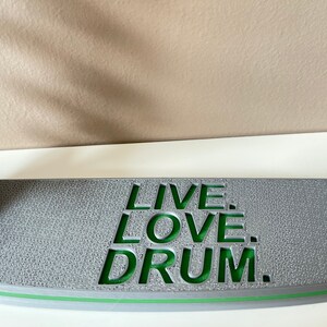 K&ouml;nnte beinhalten: Grauer Holz-Drum-St&auml;nder mit einer gr&uuml;nen Aufschrift, die "LIVE. LOVE. DRUM." lautet.