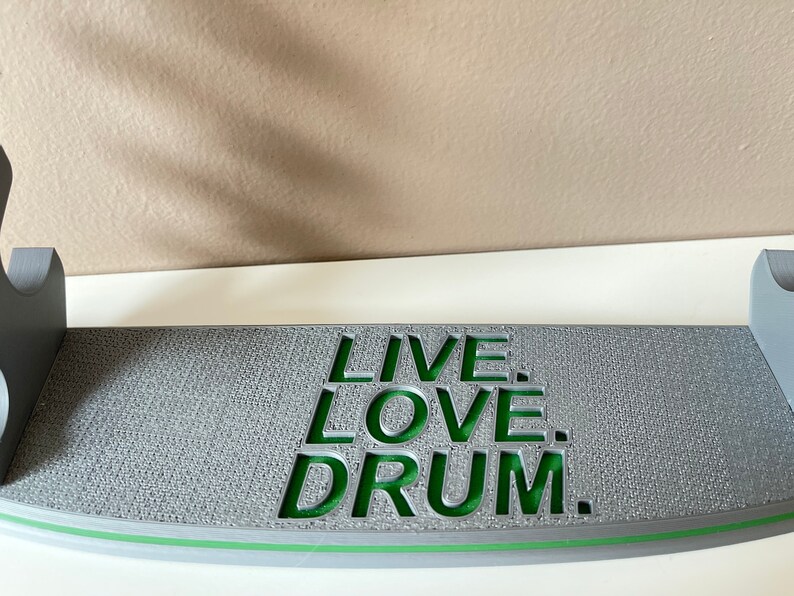 K&ouml;nnte beinhalten: Grauer Holz-Drum-St&auml;nder mit einer gr&uuml;nen Aufschrift, die "LIVE. LOVE. DRUM." lautet.