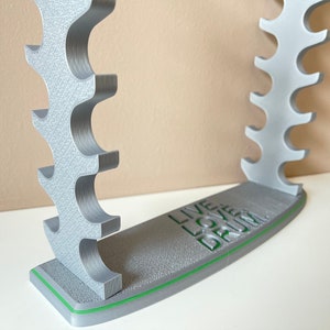K&ouml;nnte beinhalten: Ein silberfarbener, 3D-gedruckter Schlagzeugst&ouml;ckehalter mit gr&uuml;nem Sockel. Auf dem Sockel steht "Live Love Drum" gedruckt.