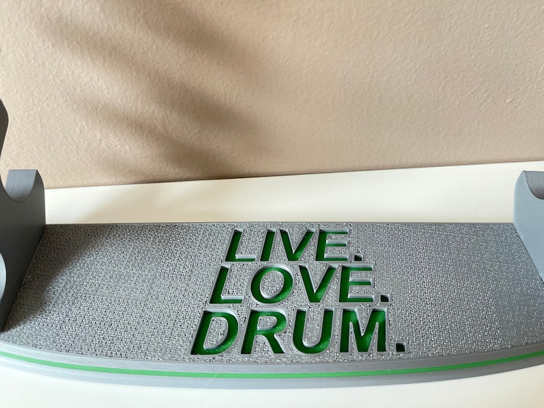 K&ouml;nnte beinhalten: Grauer Holz-Drum-St&auml;nder mit einer gr&uuml;nen Aufschrift, die "LIVE. LOVE. DRUM." lautet.