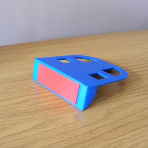 May include: A blue plastic phone stand with a red adhesive pad on the bottom. The stand has two rectangular cutouts on the top.