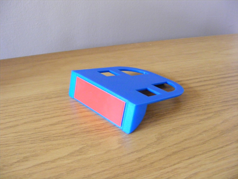 May include: A blue plastic phone stand with a red adhesive pad on the bottom. The stand has two rectangular cutouts on the top.