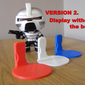 May include: A silver and black action figure stands on a white display stand. The figure is holding a gun in its right hand. The figure is standing on a wooden table. There are three display stands in the image: one red, one white, and one blue.