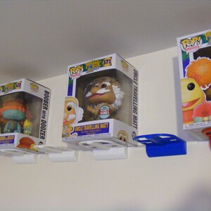May include: Three Funko Pop! vinyl figures displayed on white wall shelves with blue and red brackets. The figures are from the Fraggle Rock TV show. The figures are Boober with Doozer, Uncle Travelling Matt, and Red with Doozer.