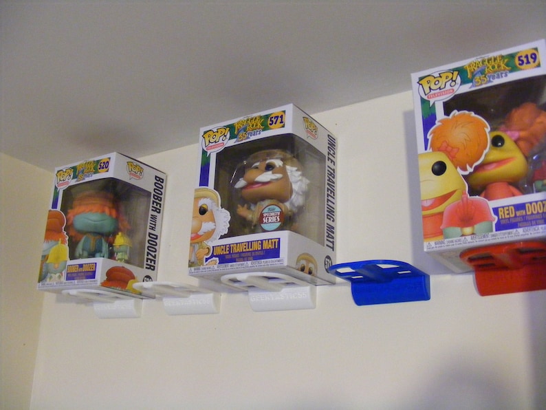 May include: Three Funko Pop! vinyl figures displayed on white wall shelves with blue and red brackets. The figures are from the Fraggle Rock TV show. The figures are Boober with Doozer, Uncle Travelling Matt, and Red with Doozer.