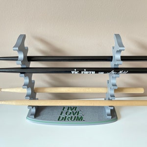 K&ouml;nnte beinhalten: Ein grauer 3D-gedruckter Schlagzeugst&ouml;ckehalter mit vier Schlitzen. Der Halter hat die Worte "Live, Love, Drum." auf der Basis aufgedruckt. Es befinden sich vier Schlagzeugst&ouml;cke im Halter, zwei schwarze und zwei aus Holz.