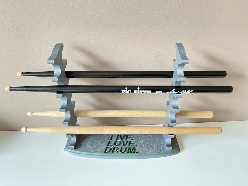 K&ouml;nnte beinhalten: Ein grauer 3D-gedruckter Schlagzeugst&ouml;ckehalter mit vier Schlitzen. Der Halter hat die Worte "Live, Love, Drum." auf der Basis aufgedruckt. Es befinden sich vier Schlagzeugst&ouml;cke im Halter, zwei schwarze und zwei aus Holz.
