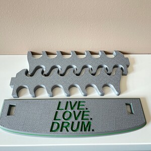 K&ouml;nnte beinhalten: Grauer 3D-gedruckter Schlagzeugst&ouml;ckehalter mit einer gebogenen Basis, auf der "LIVE. LOVE. DRUM." in gr&uuml;ner Schrift steht.