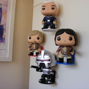 May include: Four pop culture figures are displayed on a white wall. The figures are mounted on small shelves. The figures are a silver robot, a man with blonde hair, a man with black hair, and a man with gray hair. The figures are all wearing different outfits.