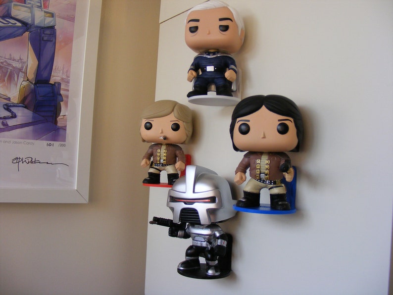 May include: Four pop culture figures are displayed on a white wall. The figures are mounted on small shelves. The figures are a silver robot, a man with blonde hair, a man with black hair, and a man with gray hair. The figures are all wearing different outfits.