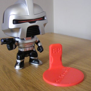 May include: A silver and black robot action figure with a gun in its hand. The figure is standing on a red plastic stand with the text "F.P. Geek Merchandise" printed on it.