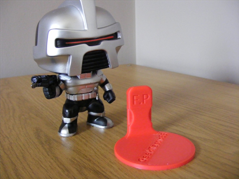 May include: A silver and black robot action figure with a gun in its hand. The figure is standing on a red plastic stand with the text "F.P. Geek Merchandise" printed on it.