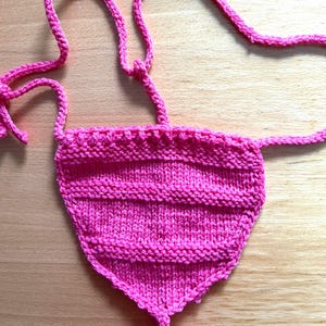Pode incluir: Uma peça de malha triangular em rosa brilhante com nervuras horizontais texturizadas e laços finos de malha que se estendem dos cantos superiores. A peça é construída com uma combinação de pontos de malha e cordões em fio vibrante.