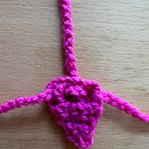 Pode incluir: Uma peça de crochê em fúcsia brilhante com formato triangular e três cordões de fio anexados que se estendem do topo e das laterais. O item é feito de uma fibra rosa vibrante com uma textura de nós visível em toda a sua estrutura.