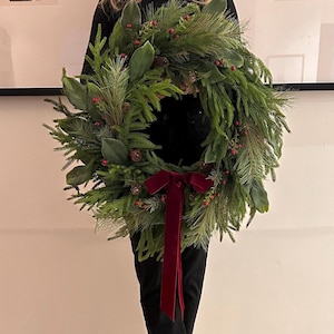 Peut inclure: Une couronne de Noël verte et luxuriante avec des aiguilles de pin, des feuilles de magnolia et de petites baies rouges. La couronne est ornée de pommes de pin et d'un nœud en velours rouge foncé. Une décoration de vacances festive.