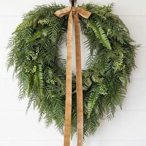 May include: A full, green wreath made from different types of leaves, including ferns and small, white berries. A wide, tan ribbon is tied at the top, with long streamers. The wreath is a decorative item, suitable for a door or wall.