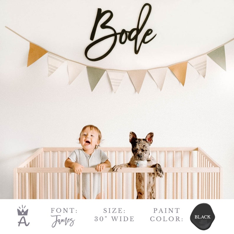 May include: Black wooden sign with the name "Bode" in a cursive font. The sign is 30 inches wide and is hanging on a white wall above a crib. A baby and a dog are standing in the crib. The sign is part of a nursery decor set.