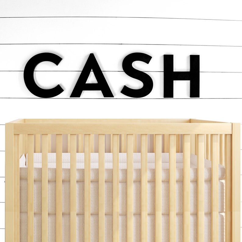 May include: Black wooden letters spelling "CASH" are mounted on a white wood wall above a light wood crib with a white mattress.