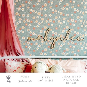 May include: A wooden sign with the name "mckynlee" in a cursive font called "sienna". The sign is 30 inches wide and is unpainted natural birch. The sign is displayed against a floral wallpaper with a light blue background and white and pink flowers.