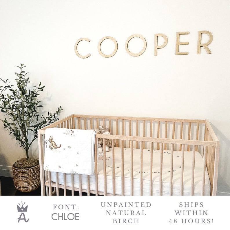 May include: Wooden nursery name sign spelling "COOPER" in a simple, modern font called "CHLOE". The sign is unpainted and made of natural birch wood. The sign is displayed above a crib with a white blanket and a stuffed animal.