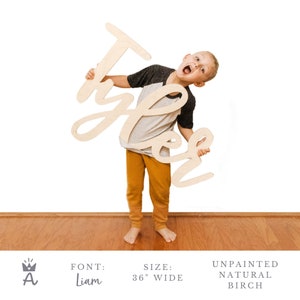 May include: A child holds up a large, wooden sign with the name "Tyler" written in a cursive font. The sign is 36 inches wide and made of natural birch wood. The sign is unpainted.