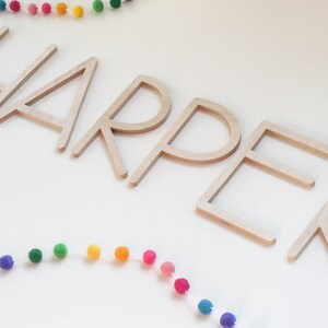 May include: Wooden letters spelling out the name "HARPER" in a simple, modern font. The letters are arranged in a line and are a light brown color. A rainbow pom-pom garland is above and below the letters.
