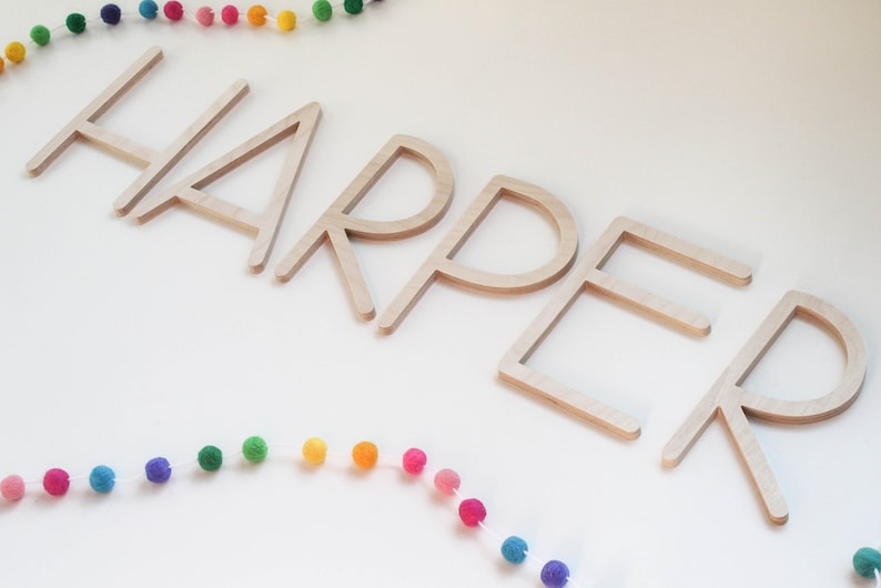 May include: Wooden letters spelling out the name "HARPER" in a simple, modern font. The letters are arranged in a line and are a light brown color. A rainbow pom-pom garland is above and below the letters.