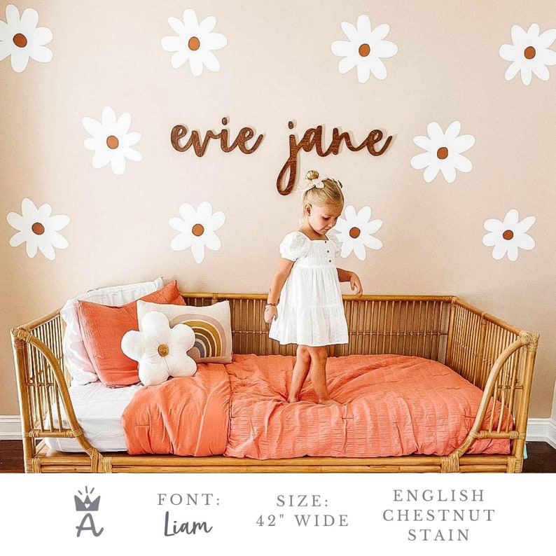 May include: A white wooden sign with the names "evie jane" written in cursive script. The sign is mounted on a light pink wall with white daisy decals. A young girl in a white dress stands in front of the sign.
