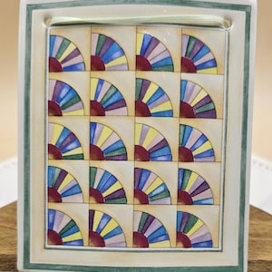 May include: A ceramic tile with a colorful geometric design. The tile features a repeating pattern of fan-shaped segments in shades of blue, green, purple, yellow, and red. The tile is framed with a green border.