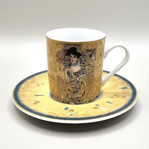May include: A gold-toned teacup and saucer set featuring a portrait of a woman in a decorative dress. The cup and saucer have a gold background with a blue border. The teacup has a white handle and interior.