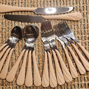 May include: A collection of stainless steel flatware with cream-colored handles. The set includes knives, forks, and spoons, arranged on a woven surface. The knives have a brand name on the blade. The handles feature a decorative design.
