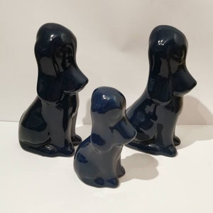 Könnte beinhalten: Drei marineblaue Keramik-Hundefiguren. Zwei sind größer und eine kleiner, alle in sitzender Position. Die glänzende Oberfläche reflektiert das Licht und hebt die glatten Kurven der Hundeskulpturen hervor. Die Figuren sind auf einer weißen Oberfläche angeordnet.