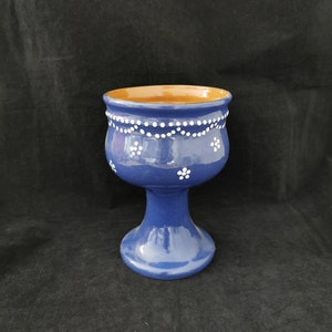 Op de afbeelding: Een blauwe keramische kelk met een wit bloemenmotief en een bruine rand. De kelk heeft een brede voet en een lange, slanke steel.