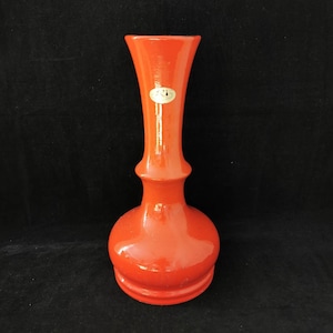 以下が含まれることがあります： 球根状のベース、細い首、フレアトップを備えた鮮やかなオレンジ色の花瓶。 花瓶は光沢のある仕上げで、上部に小さな楕円形のステッカーが付いています。 背景は無地の黒です。