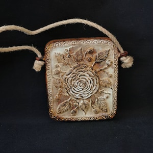 Könnte beinhalten: Ein Keramik-Wandbehang mit einer Schlaufe aus Seil zum Aufhängen. Der Wandbehang ist rechteckig und zeigt ein erhabenes Design einer weißen Rose mit Blättern und Beeren. Der Hintergrund des Wandbehangs ist hellbraun.