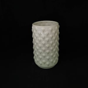 May include: A white ceramic vase with a textured surface of small, rounded, heart-shaped bumps. The cylindrical vase has a smooth, open top and sits against a black background. The vase is a decorative item.