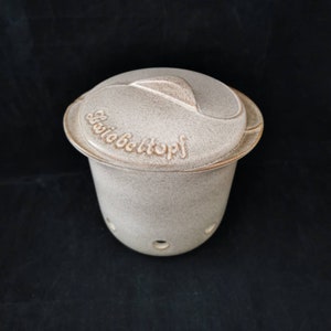 May include: A beige ceramic container with a lid and holes in the bottom. The lid has a raised handle and the words "Gießbehältnis" are embossed on the lid.