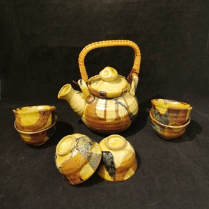 Puede incluir: Juego de té de cerámica con tetera y cuatro tazas. La tetera tiene un asa tejida y una tapa. El juego está esmaltado en tonos marrones, amarillos y negros. Las tazas están apiladas en pares.