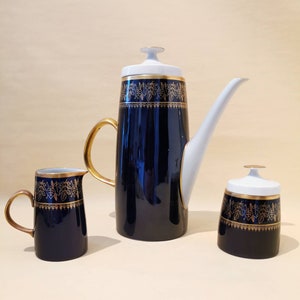 Op de afbeelding: Een vintage koffieset van drie delen. De set bestaat uit een hoge, marineblauwe koffiepot met een witte deksel en tuit, een melkkan en een suikerpot. Elk stuk is versierd met gouden bladpatronen en gouden randen.