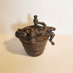 May include: A small, antique, brass container with a hinged lid and a decorative handle. The container has a cylindrical shape and a textured surface.