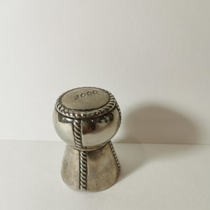 Könnte beinhalten: Silberner Champagnerflaschenverschluss mit einem Seil-Design und dem Jahr 2000, das auf der Oberseite eingraviert ist.