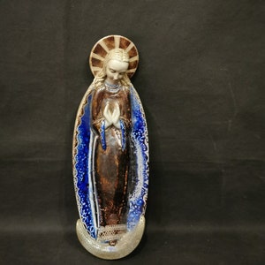 May include: A ceramic figurine of the Virgin Mary, standing in a blue and white robe with a halo. The figurine is brown and has a detailed face.
