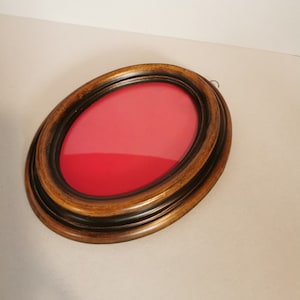 Puede incluir: Un marco de fotos ovalado de madera con un acabado marrón oscuro y un paspartú de terciopelo rojo. El marco tiene un diseño sencillo con un borde ligeramente elevado.