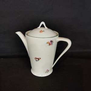Puede incluir: Tetera de porcelana blanca con tapa y asa. La tetera está decorada con pequeños motivos florales en rojo, azul y amarillo. Un ribete verde acentúa la tapa y el borde de la tetera.