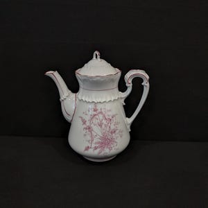 Könnte beinhalten: Weiße Porzellan-Teekanne mit einem rosa Blumendesign. Die Teekanne hat einen gebogenen Ausguss, einen dekorativen Griff und einen Deckel mit einem kleinen Knopf. Der Rand und die Kanten sind mit einem rosa Besatz versehen.