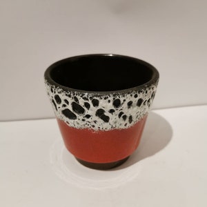 Peut inclure: Une petite tasse en céramique avec un intérieur noir et un extérieur rouge. La partie supérieure de la tasse présente un motif tacheté blanc et noir. La tasse a une forme arrondie et repose sur une petite base marron foncé.