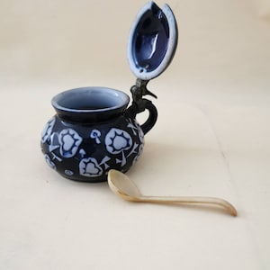 Può includere: Un contenitore in ceramica blu scuro con coperchio incernierato e manico piccolo. Il contenitore è decorato con motivi floreali bianchi. Un piccolo cucchiaio colorato chiaro è appoggiato accanto.