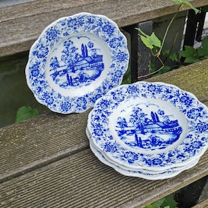 May include: A collection of white ceramic plates featuring a blue floral and landscape pattern. The plates have a scalloped edge and are arranged on a wooden surface. The landscape design shows a building, trees, and water.