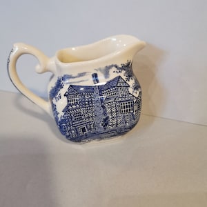 Può includere: Un brocca per la panna in ceramica bianca con un motivo blu e bianco di una casa e alberi. La brocca ha un manico e un beccuccio.